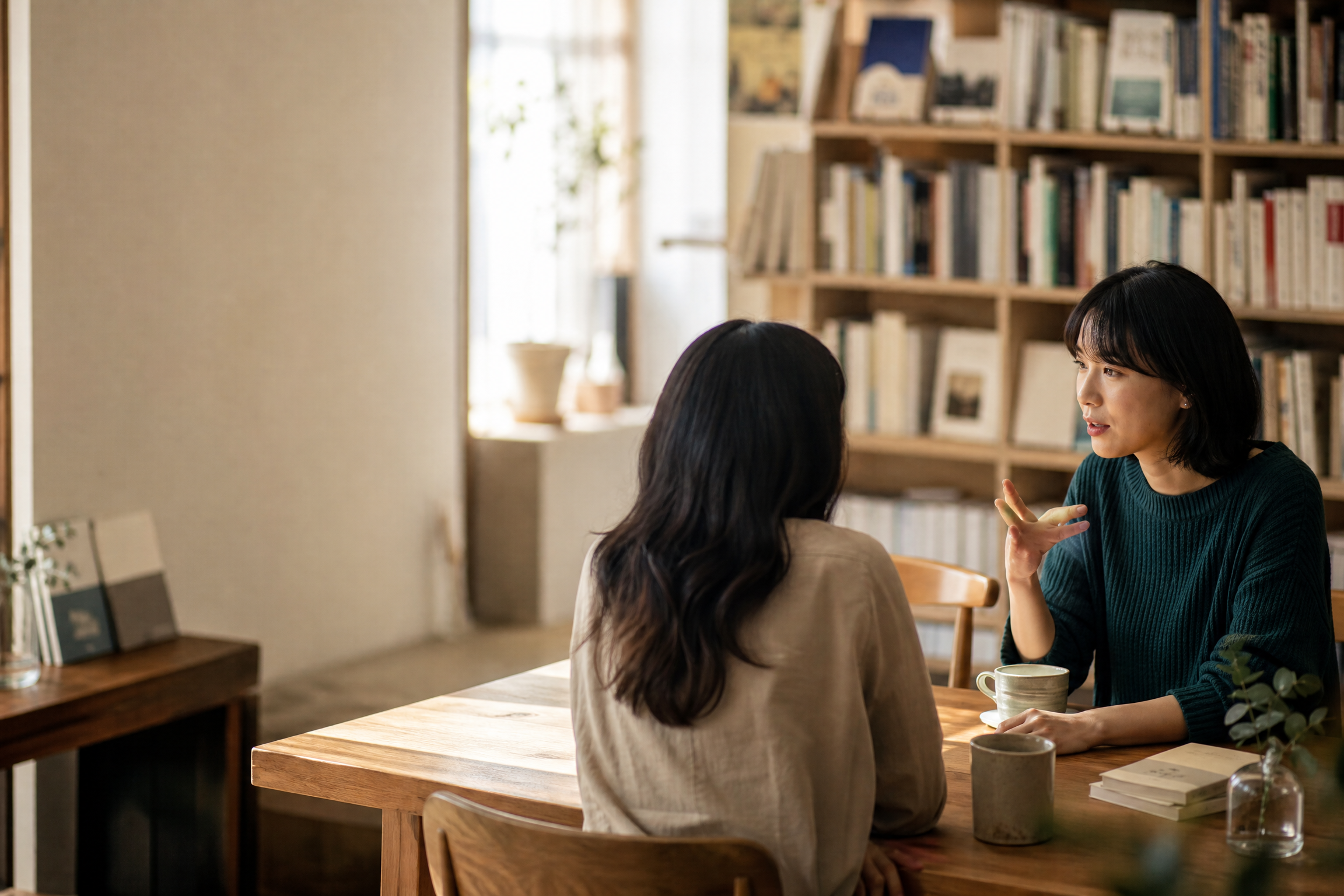 Two people talking across a wooden table in a sunlit room with bookshelves.